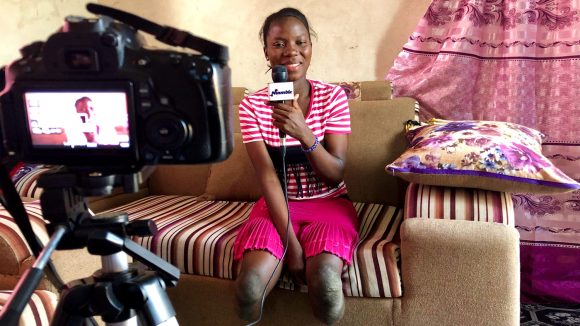 A woman with amputated legs sits on a sofa, smiling and speaking into a microphone in front of a camera.