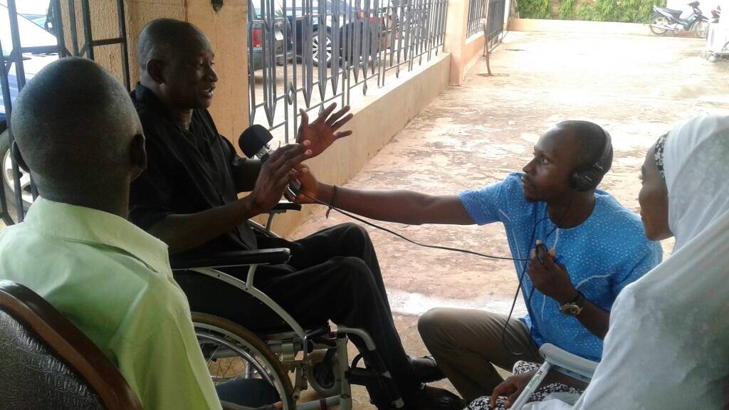 A man in a wheelchair is interviewed by a man holding a microphone and wearing headphones.