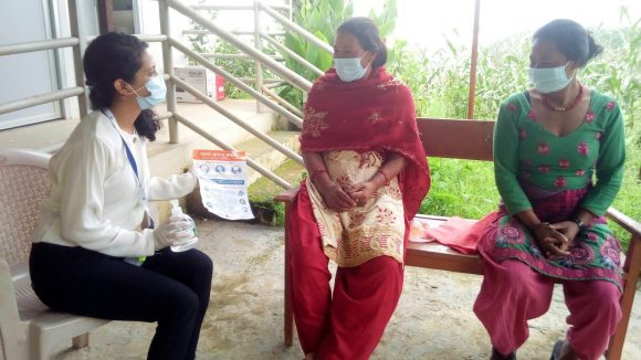 Three women sit talking in Nepal. They are wearing facemasks and one is holding a bottle of sanitiser and an information leaflet.