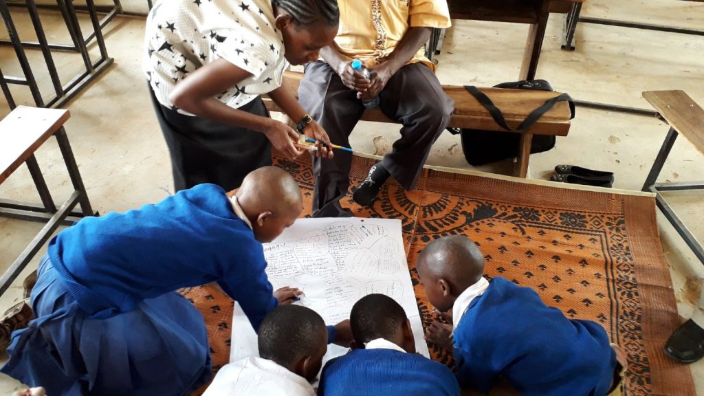 Peer research by Institute of development studies in Tanzania. Four children in school uniform sit on the floor and are writing on a large piece of paper, supervised by two adults.