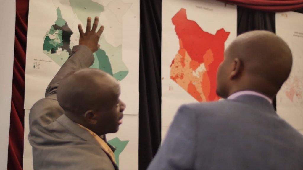 Two men talk together in front of illustrated maps.