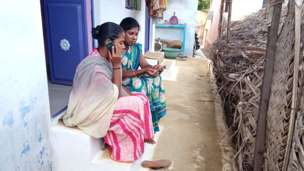 Two women sit on a doorstep, one is speaking into a mobile phone.