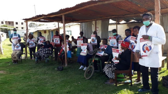 A group of seventeen people from an organisation of people with disabilities smiling at the camera and sitting under a wooden structure. They are holding posters.