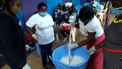 Four women are stood around a blue bucket and wearing masks. One of the women is stirring a white liquid in the bucket.