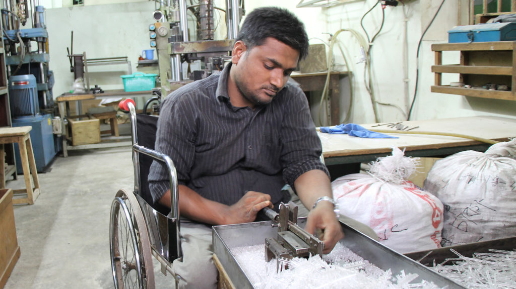 A man using a wheelchair operates a small machine in a factory to make plastic manufacturing pieces.