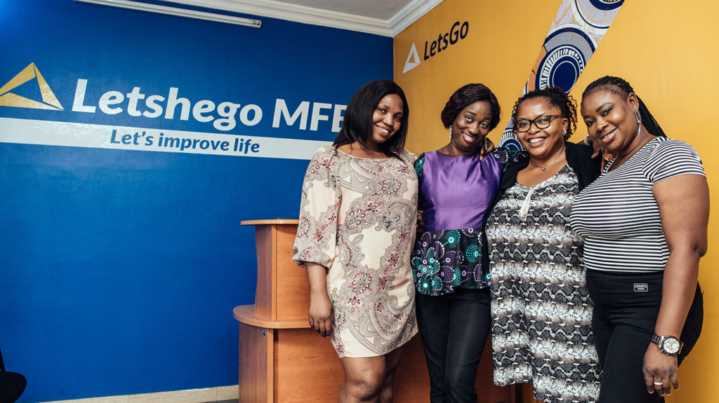 Four women smile while standing ni the reception area of Letshego Microfinance Bank, with branded logos on the blue and yellow walls behind them.