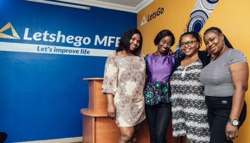 Four women smile while standing ni the reception area of Letshego Microfinance Bank, with branded logos on the blue and yellow walls behind them.