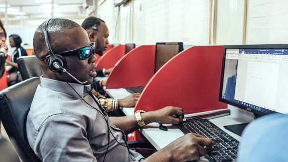 A staff member at a call centre wears a headset and sits at a computer. He's wearing dark glasses and smart clothes.