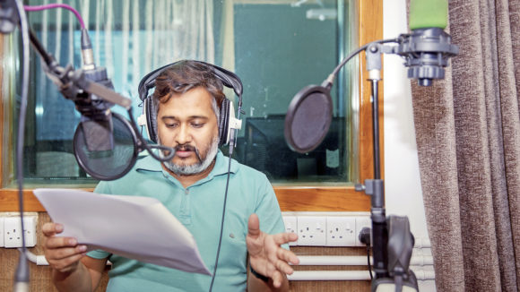 A journalist sits in a radio studio holding a script and talking into a microphone.