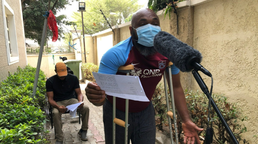 A black man with crutches reads from a script and speaks into a microphone.