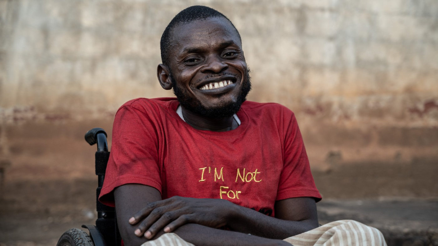 A black man in a wheelchair wearing stripy trousers and a red T-shirt smiles broadly at the camera