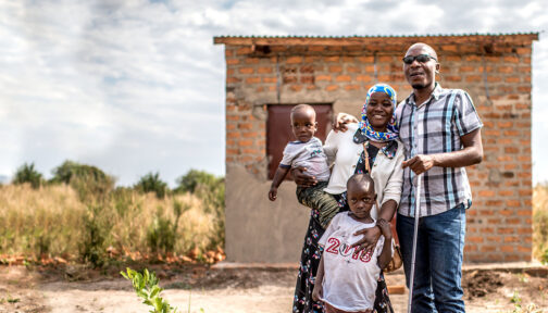 Julius and Najiba stand outside their home in Uganda with their two young children. Julius is holding a white cane.