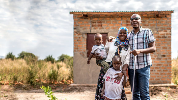 Julius and Najiba stand outside their home in Uganda with their two young children. Julius is holding a white cane.