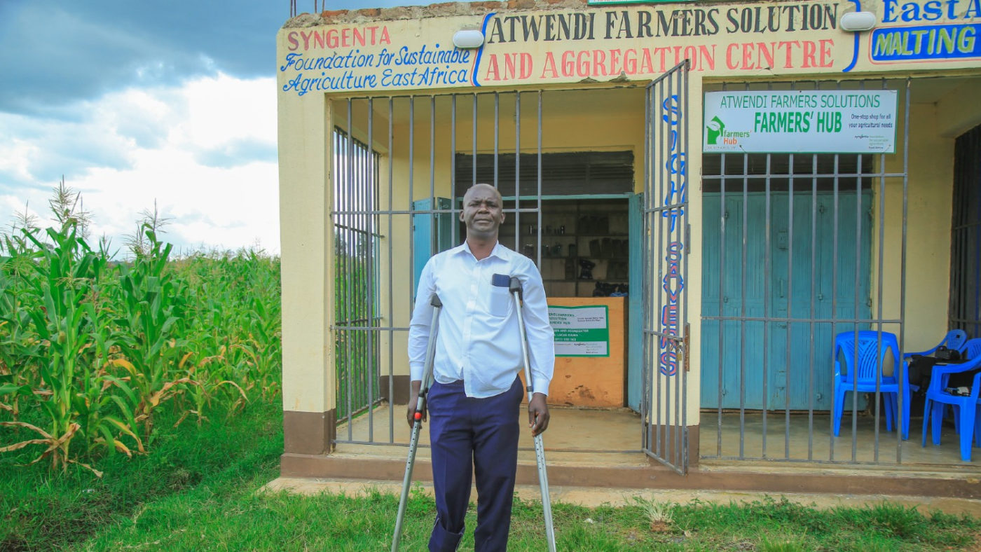 Lucas, a farmer with a disability and a network hub manager stands in front of his shop. The writing on the shop says: “Syngenta Foundation for Sustainable Agriculture East Africa. Atwendi Farmers Solution and Aggregation Centre.”