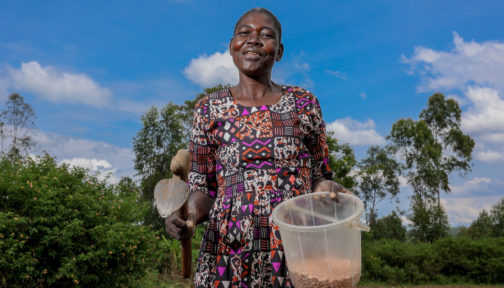 A woman farmer is photographed on her land holding farming tools and seeds.