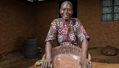 Jane Akinyi is smiling at the camera and holding a basket of seeds.