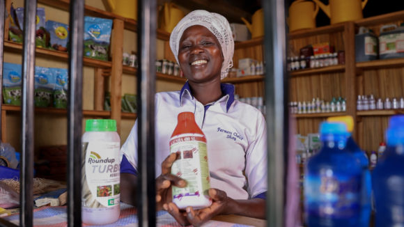 Linet Lucy is photographed in her shop. She is smiling at the camera and holding some of the farm products she sells.