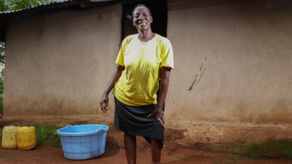 Maren Atieno, a sorghum farmer stands outside her home. She is smiling.