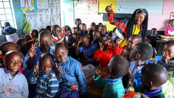 A group of school children smile at the camera inside their classroom.