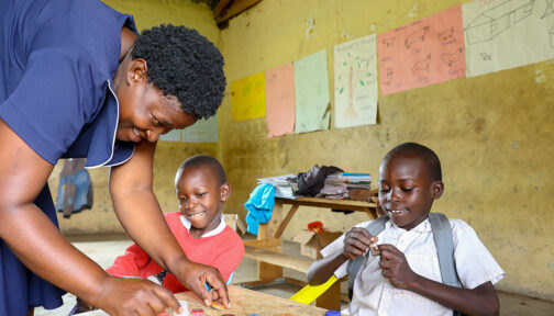A teacher in a classroom in Kenya leans down to teach two boys a task with bottle tops.