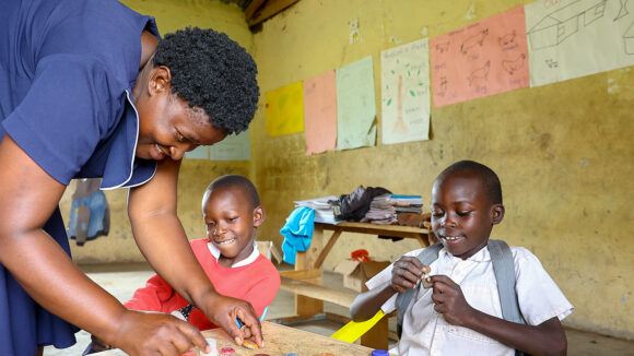 A teacher in a classroom in Kenya leans down to teach two boys a task with bottle tops.