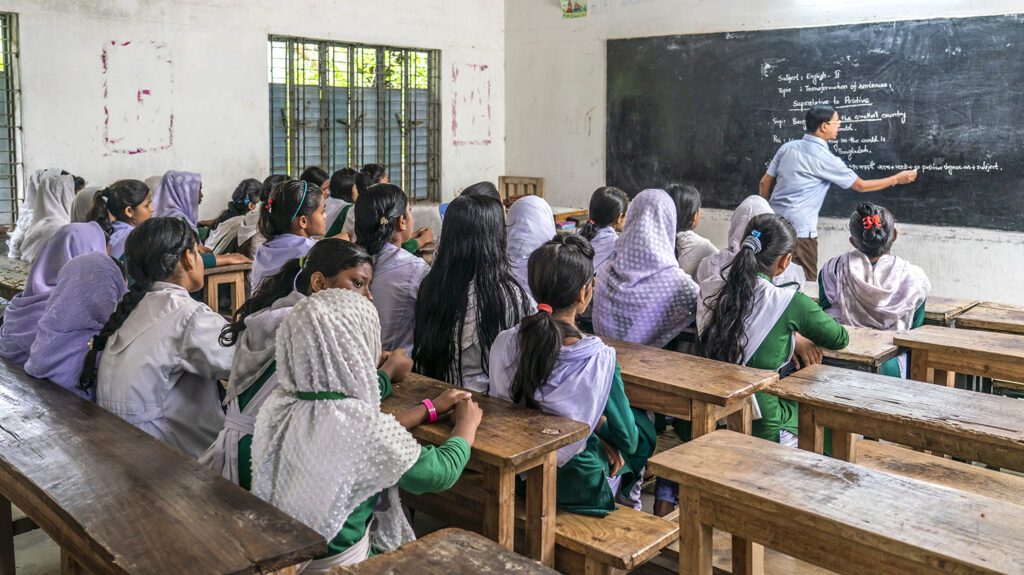 A group of school students in Bangladesh look at the blackboard in the classroom as their teacher writes on the board.