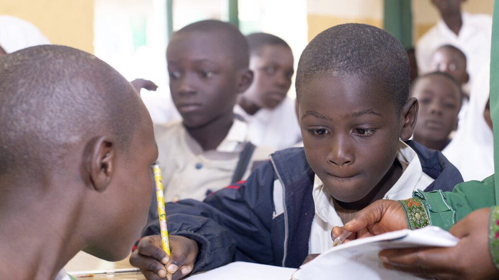 A young student in a classroom in Nigeria, holding a pencil and writing on a pad of paper.