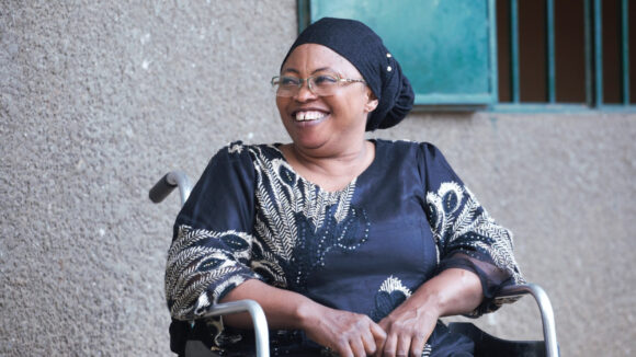A woman in a wheelchair wearing a black headscarf and dress, smiles at the camera.