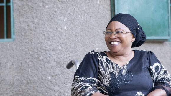 A woman in a wheelchair wearing a black headscarf and dress, smiles at the camera.