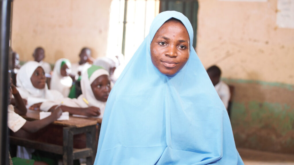 A female teacher looks into the camera as her pupils study in class behind her.