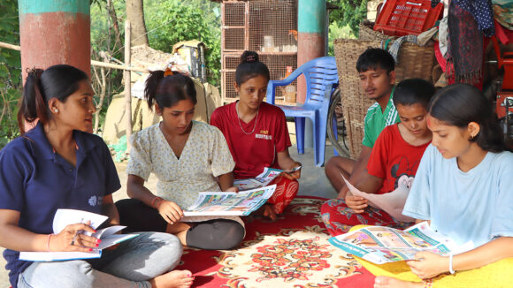 A group of women in Nepal attending an inclusive sexual health group. They're sitting on colourful rugs on the floor and looking at leaflets.