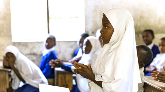 A female student in Tanzania stands and speaks in the classroom, wearing a white headscarf.