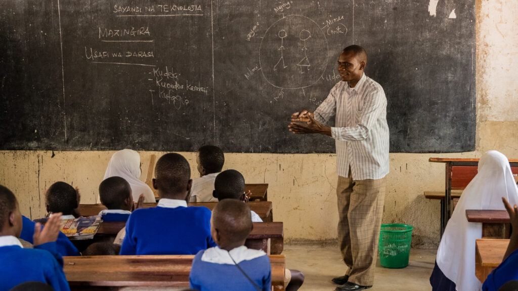 A man stands at the front of the classroom next to a blackboard. He is teaching a class of primary school-age boys and girls.