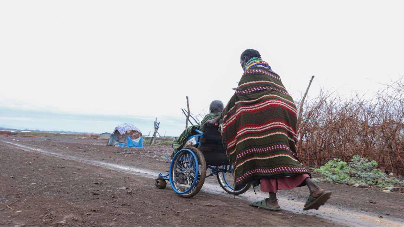 A woman pushing a wheelchair with a child on a muddy road in Kakuma refugee camp.