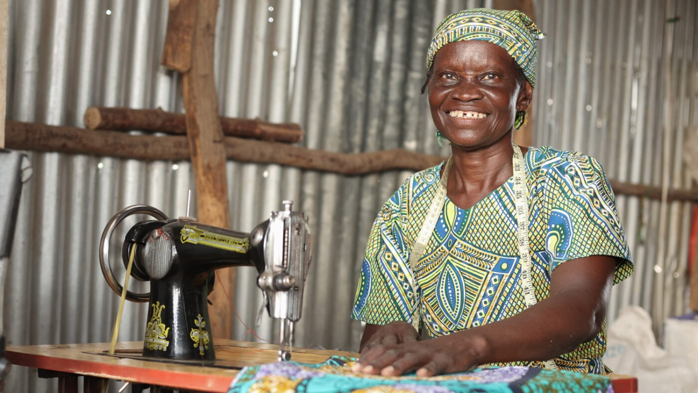 A woman wearing a colourful dress and a headscarf, sitting inside a room with her sewing machine, smiling at the camera