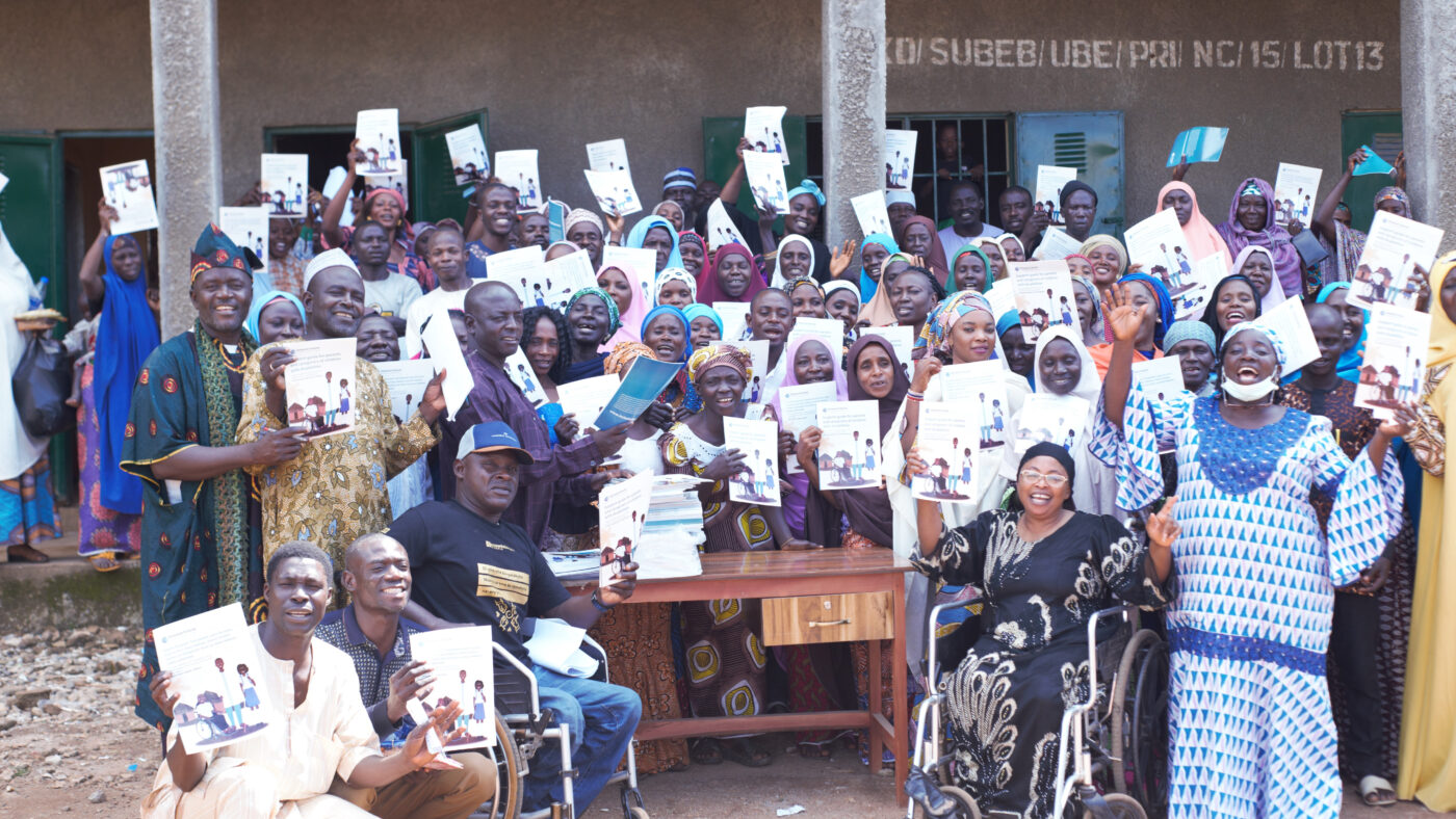 A group of people are smiling and holding up A4 guides, outside a school in Nigeria.
