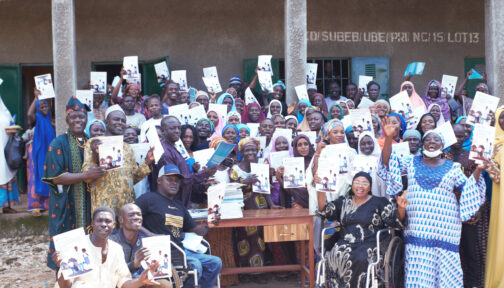 A group of people are smiling and holding up A4 guides, outside a school in Nigeria.