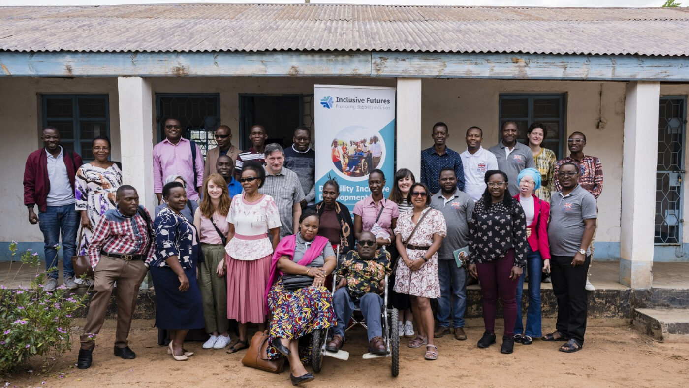 A group of men and women, with and without disabilities are stood outside of a white, single-story building. There is a banner behind them showing the words 'Inclusive Futures.'