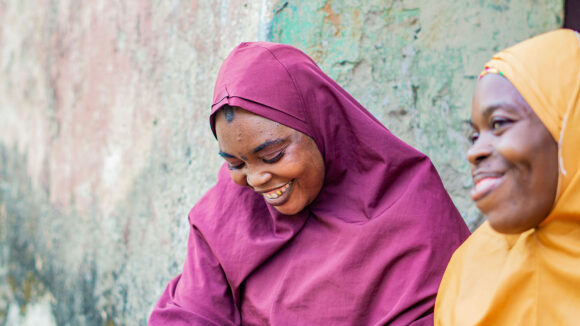 Two women wearing plum and yellow-coloured dresses and headscarfs are smiling and laughing.