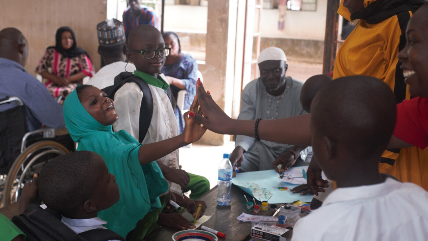 A group of children are sat and stood around a table during a school painting activity. One of the girls, wearing a blue headscarf is giving a high-five to a woman across the table.