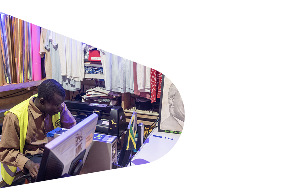 Titus sits at a desk, working at a computer. Around him are t-shirts, posters and flags.