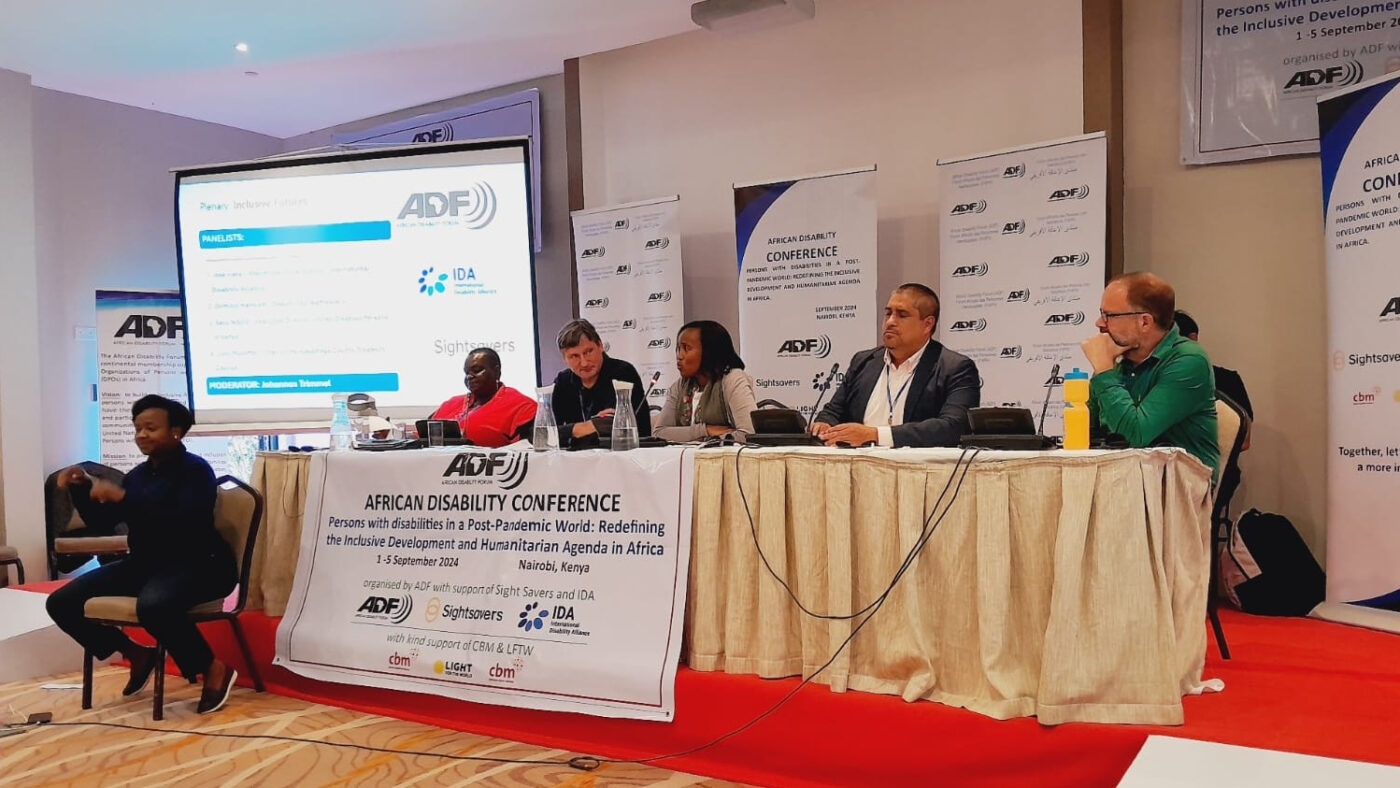 Two black women and three white men are sat at a long table - presenting as part of a panel. There are banners and a screen behind them saying 'African Disability Forum.' There is also a sign language interpreter to the left of the panel.