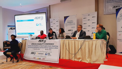 Two black women and three white men are sat at a long table - presenting as part of a panel. There are banners and a screen behind them saying 'African Disability Forum.' There is also a sign language interpreter to the left of the panel.