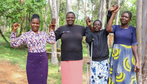 Four female sorghum farmers hold each others hands in the air while smiling. One of the women is using a crutch for assistance.