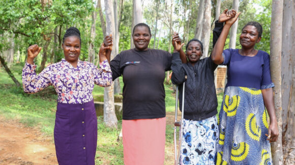 Four female sorghum farmers hold each others hands in the air while smiling. One of the women is using a crutch for assistance.