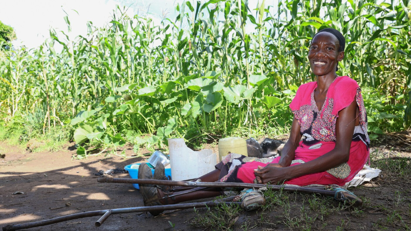 Eunice, who has a physical impairment, sits on the ground in front of a sorghum field. A pair of crutches lie next to her.