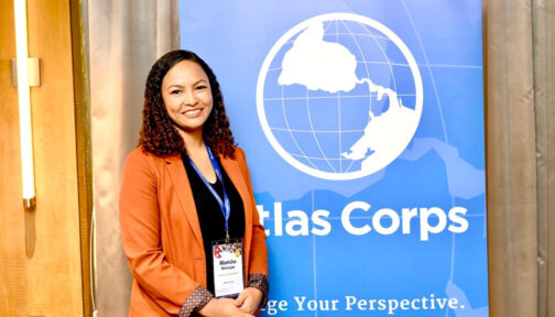 Manisha is wearing an orange jacket and smiling at the camera. Behind her is a banner saying 'Atlas Corps.'