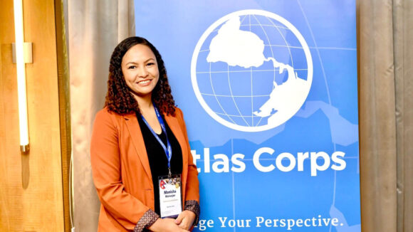 Manisha is wearing an orange jacket and smiling at the camera. Behind her is a banner saying 'Atlas Corps.'