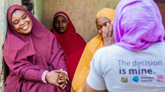 Two women wearing plum and yellow-coloured dresses and headscarfs are smiling at a woman who is wearing a T-shirt saying: 'The decision is mine.'