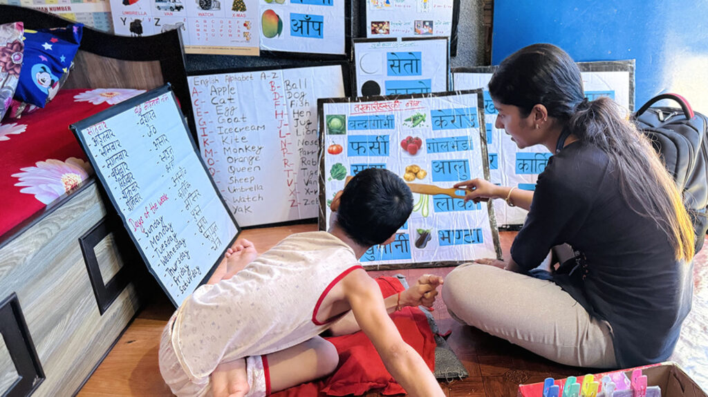 A female teacher points to a board with images of vegetables alongside the words for them. A young girl with physical disabilities who is sitting on a cushion next to her watches.